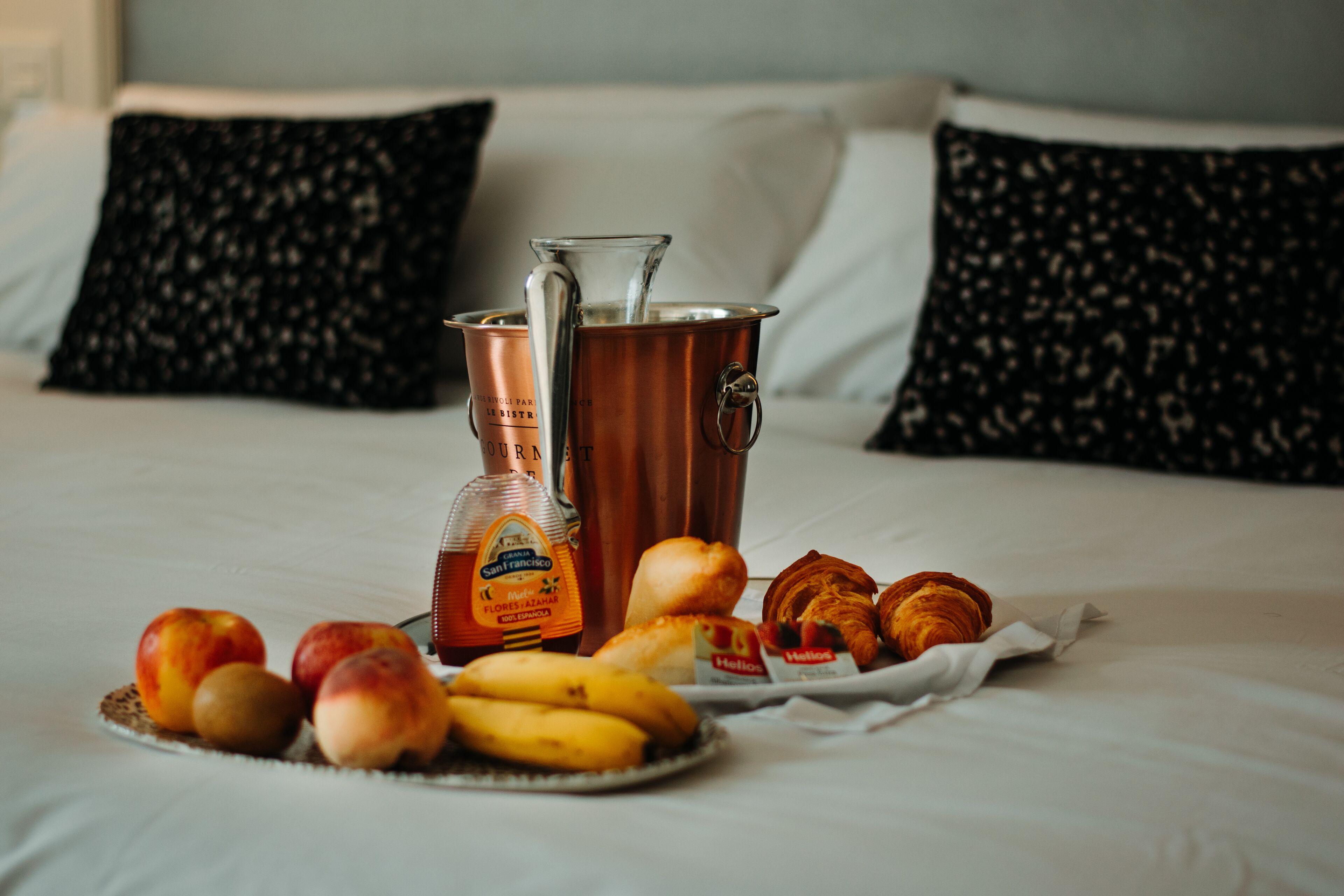 A bed with a tray of fruit, a bottle of olive oil, and a copper pot on a hotel bed.