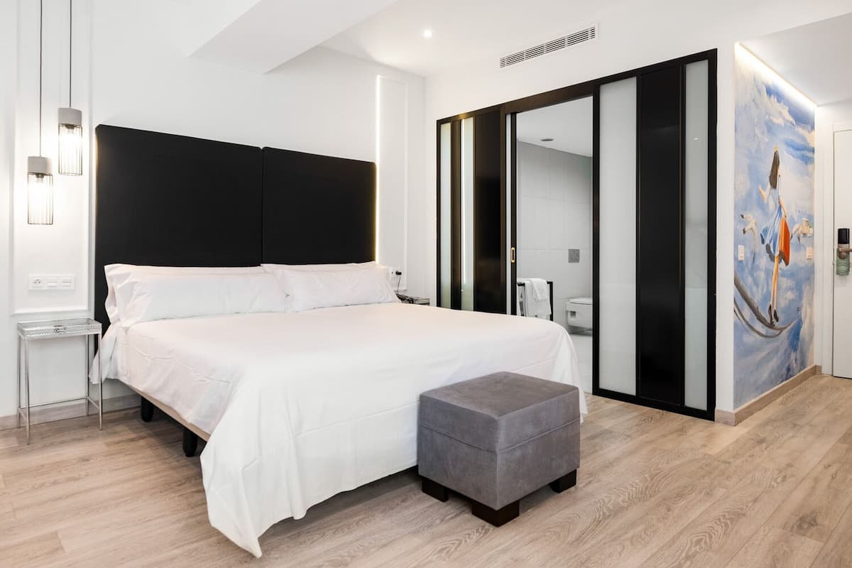 Un dormitorio moderno con una cama grande, un banco gris, un armario con espejo y un cuadro en la pared.
