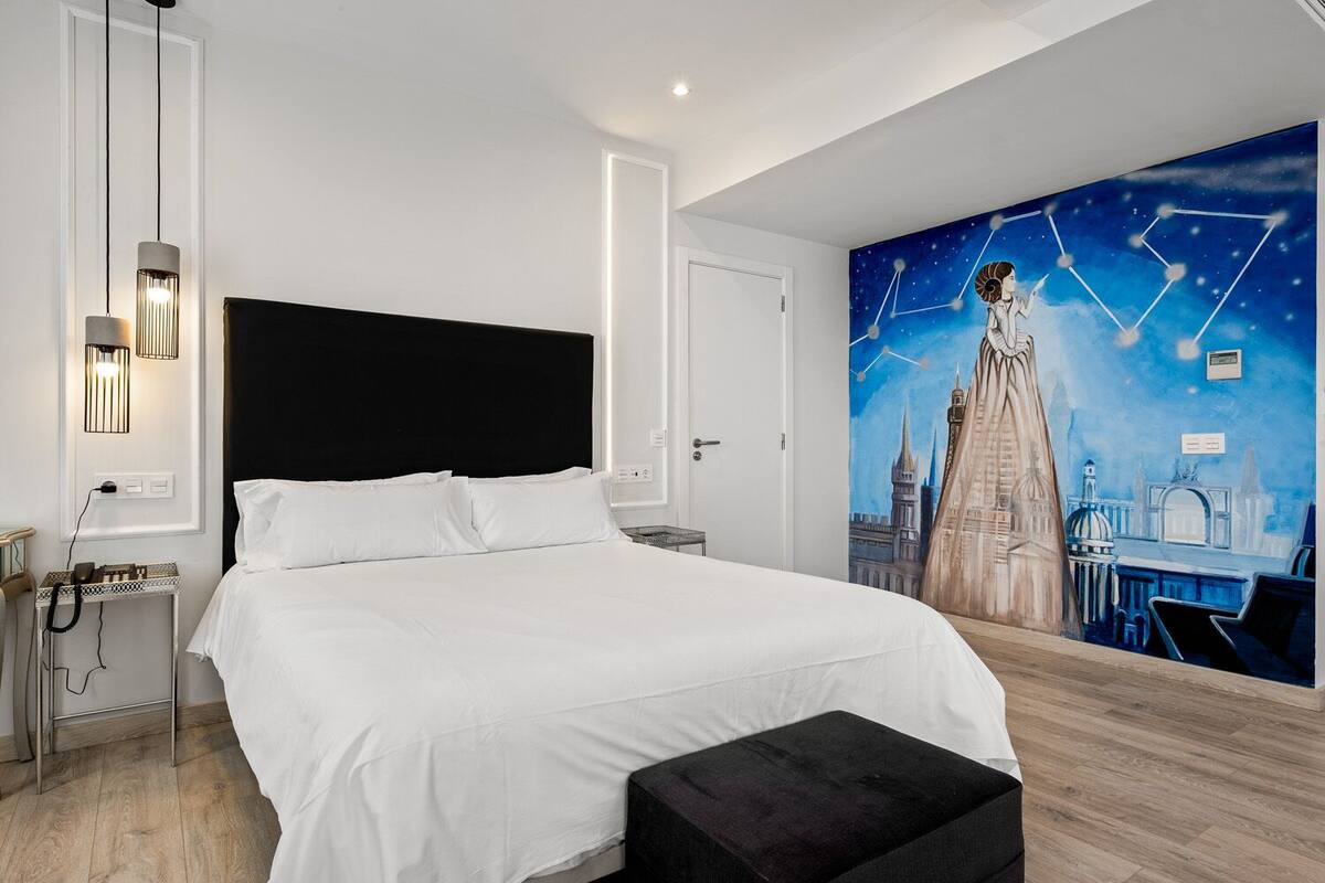 Una habitación de hotel moderna con una cama grande, un cabecero negro, un mural de un paisaje urbano y una pequeña mesa con una lámpara.