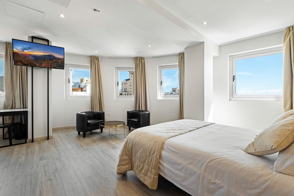 Une chambre d’hôtel moderne dotée d’un grand lit, d’un bureau avec une chaise, d’une petite table et de deux fenêtres donnant sur la ville.