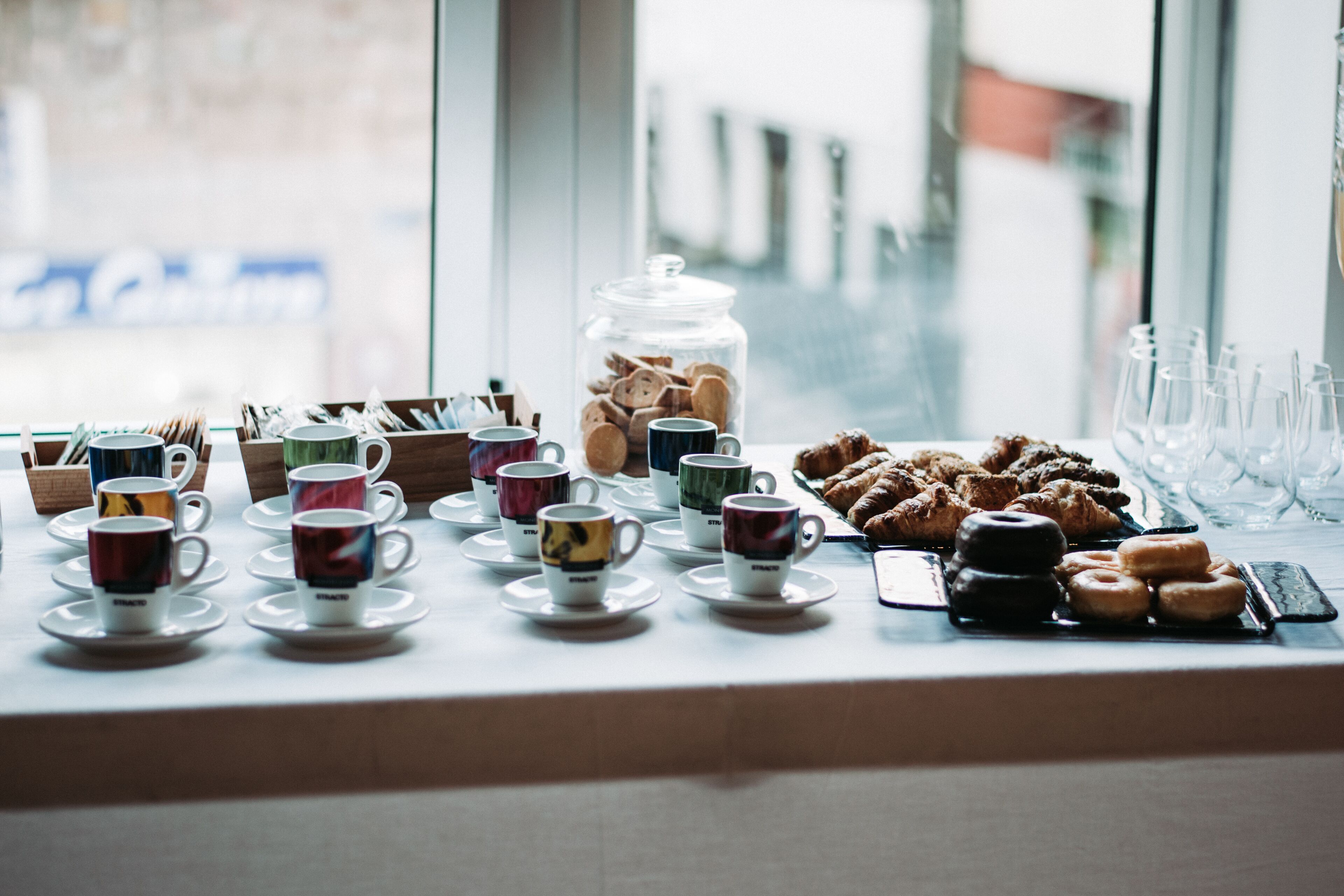A breakfast setup with various cups, pastries, and a jar of cookies on a table by a window.