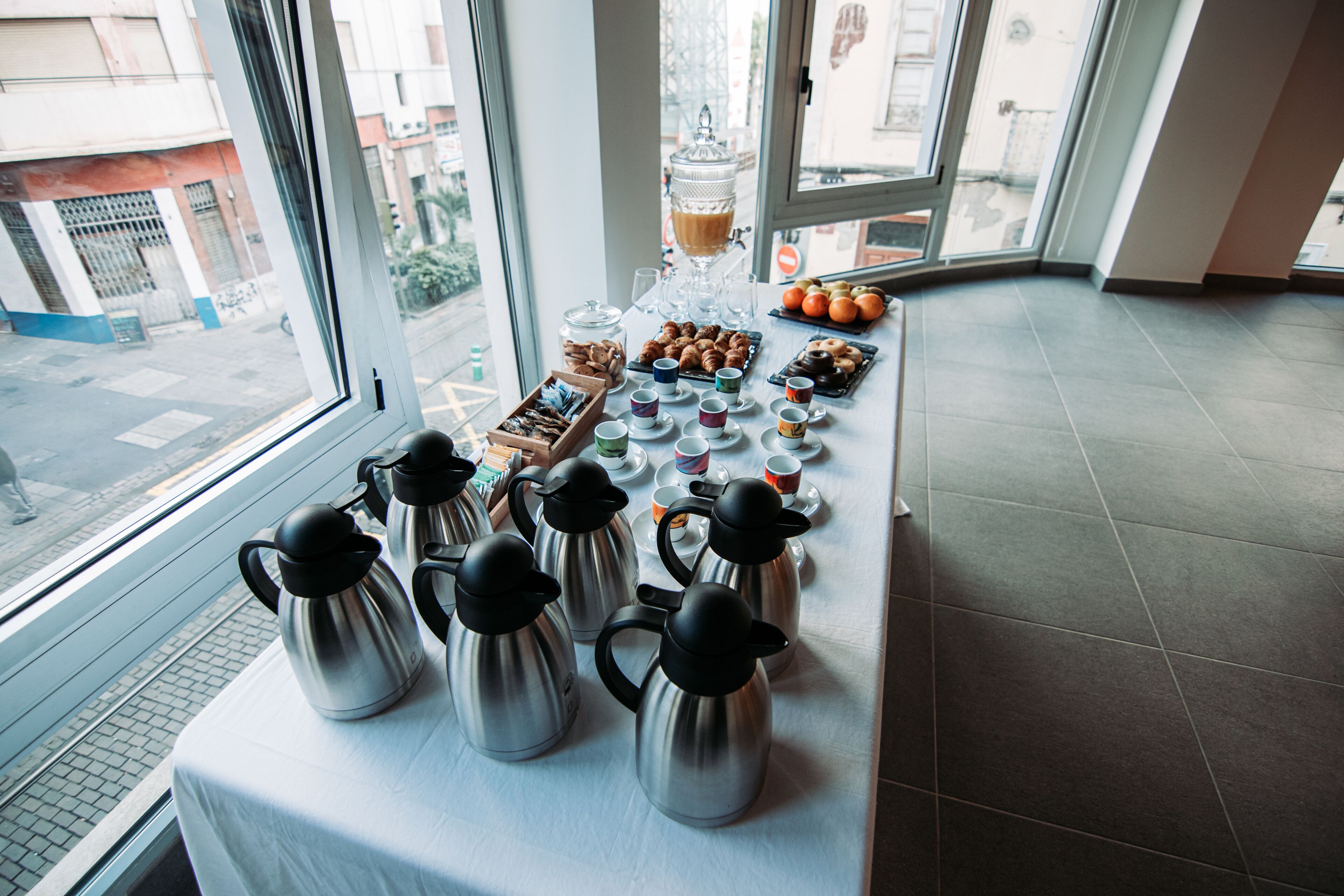 A breakfast buffet with coffee pots, pastries, and fruit on a table by a window.