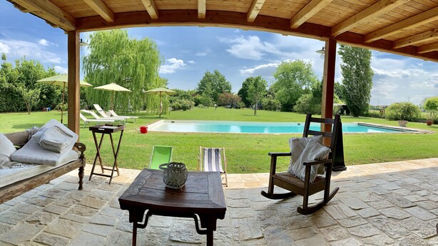 Un porche cubierto con suelo de piedra, una mesa de madera, un sofá y dos mecedoras. Se observa una piscina al fondo.