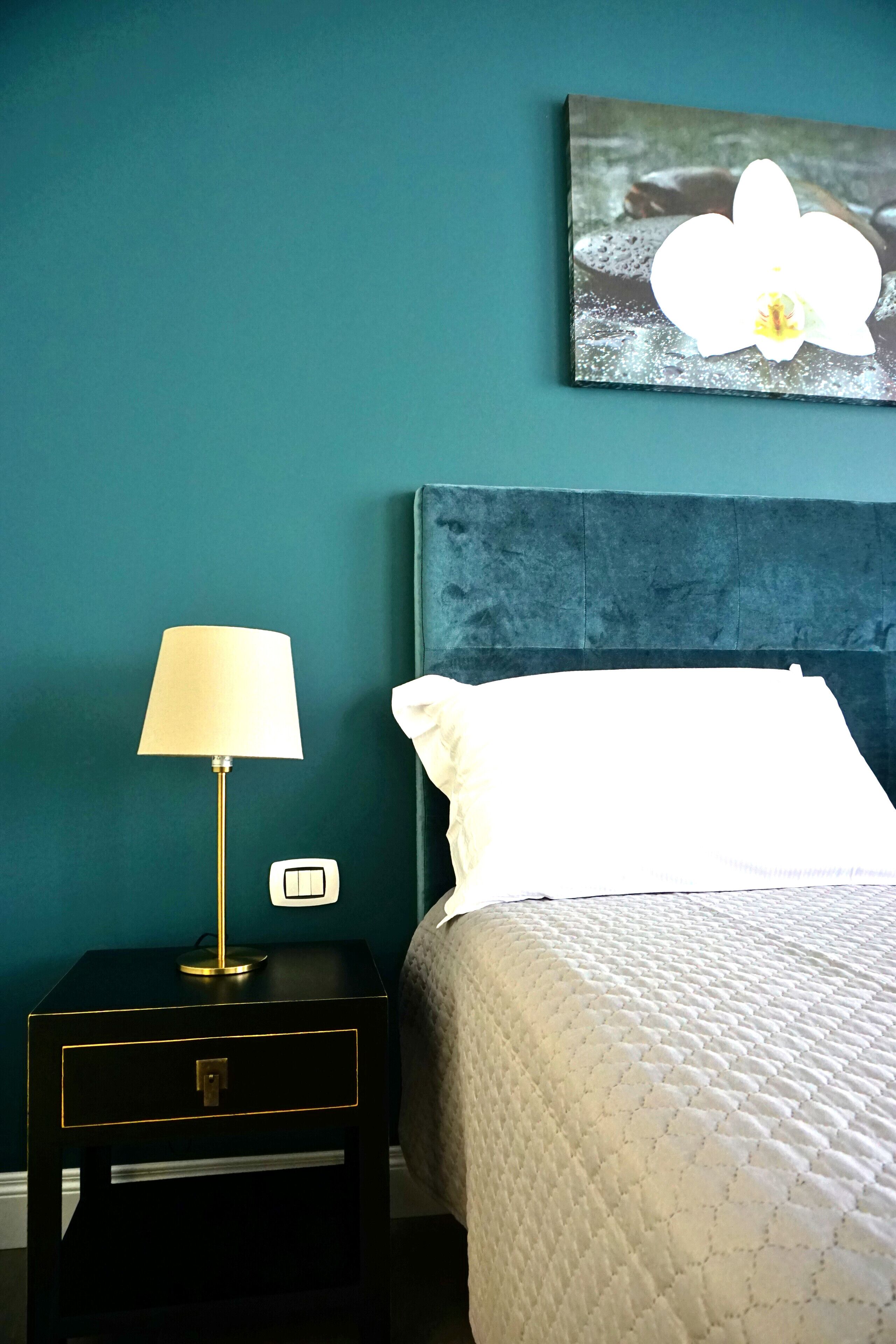 Un dormitorio con paredes color turquesa, una cama con ropa blanca, una mesita de noche con una lámpara y un cuadro de una orquídea.