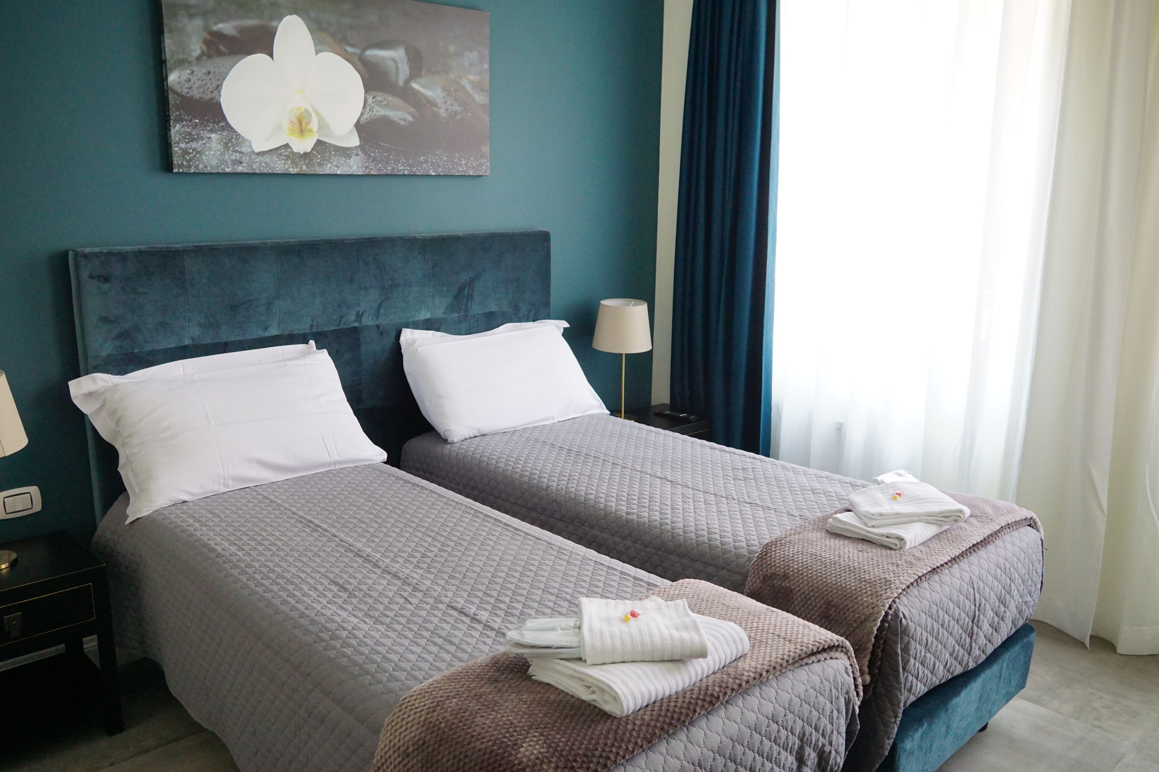 A hotel room with two beds, each with a grey quilted bedspread and white pillows. There is a painting of an orchid on the wall, a lamp on the nightstand, and a window with blue curtains.