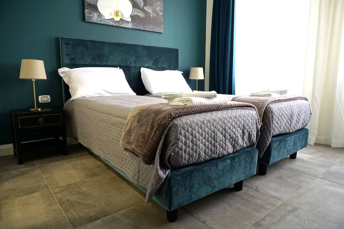 A neatly made double bed with a quilted bedspread and two pillows, flanked by bedside tables with lamps, in a room with teal walls and a large window with curtains.