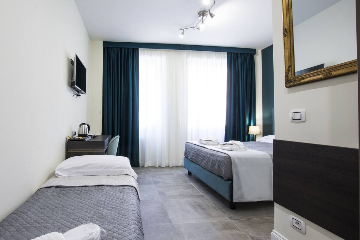 A hotel room with two single beds, a desk with a chair, a flat-screen TV, a ceiling light, and a large mirror.
