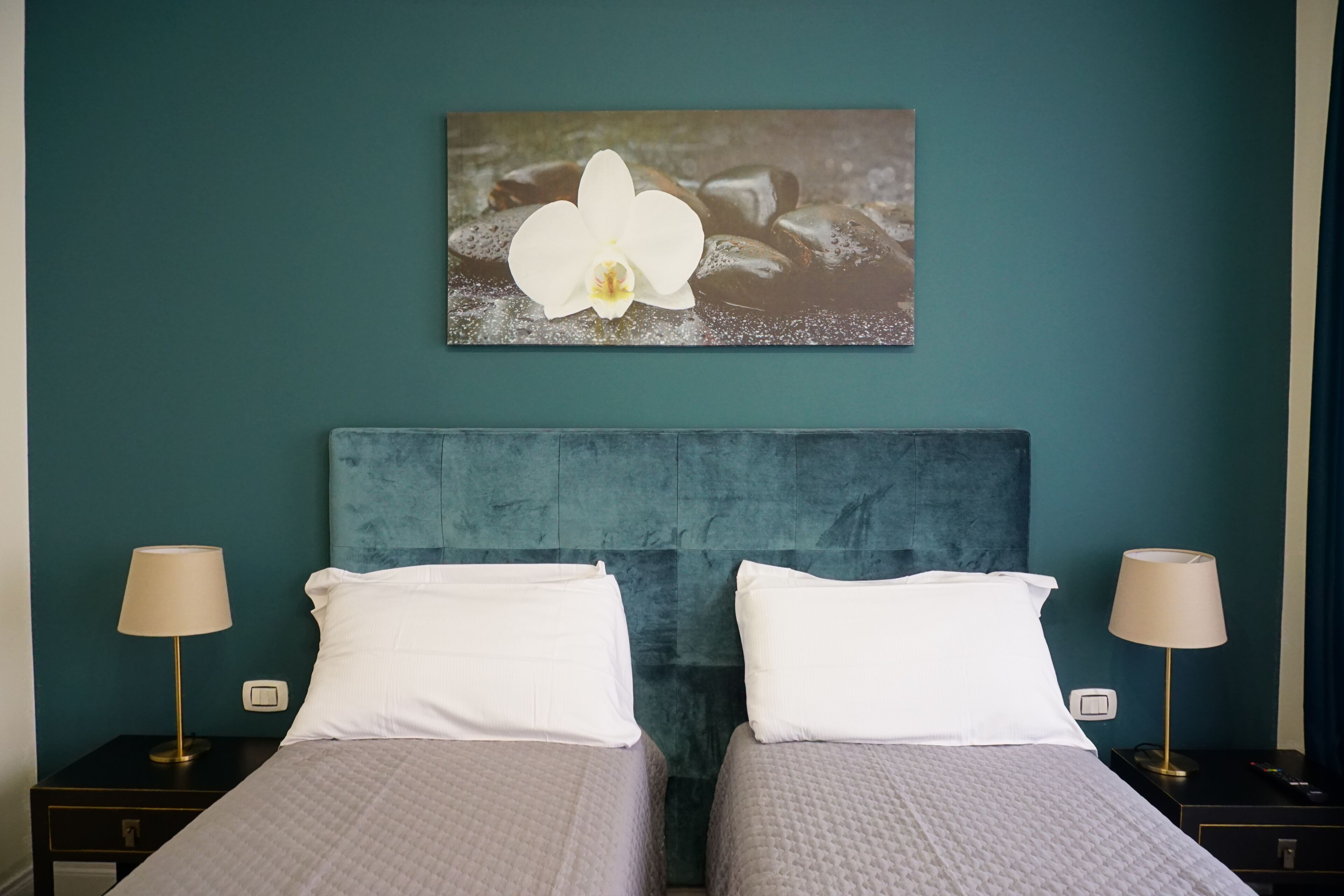 Une chambre d’hôtel aux murs de couleur turquoise, ornée d’un grand tableau représentant une orchidée, avec deux lits dotés d’oreillers blancs et de lampes de chevet.