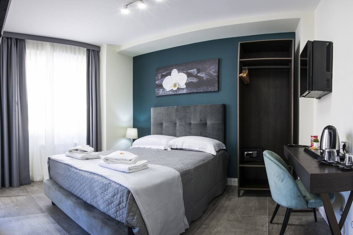 A modern hotel room with a large bed, a desk with a chair, a wardrobe, and a painting on the wall.