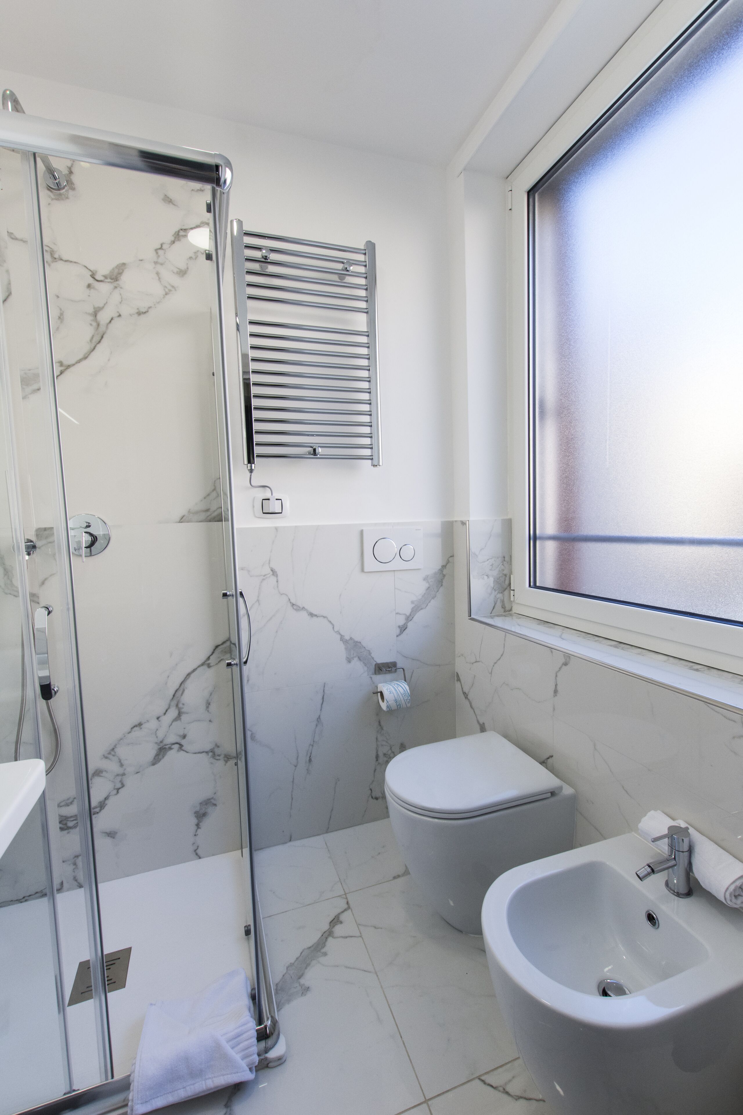 A bathroom with a glass shower enclosure, a bidet, a sink, and a window.