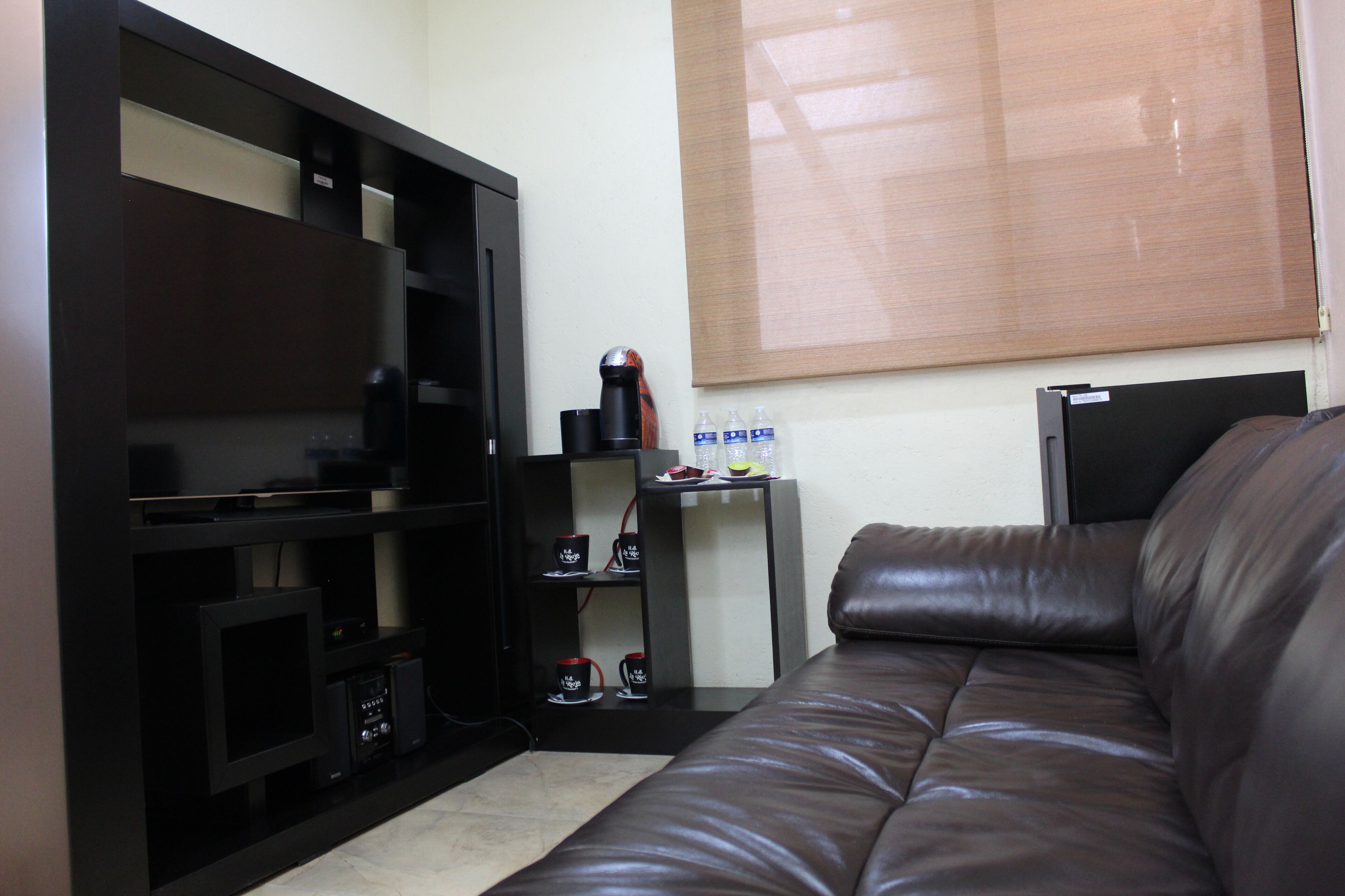 A living room with a brown leather sofa, a black entertainment unit with a flat-screen TV, a small table with a coffee maker and water bottles, and a window with blinds.