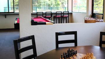 A room with a pool table, a chessboard, and a window with a view of greenery.