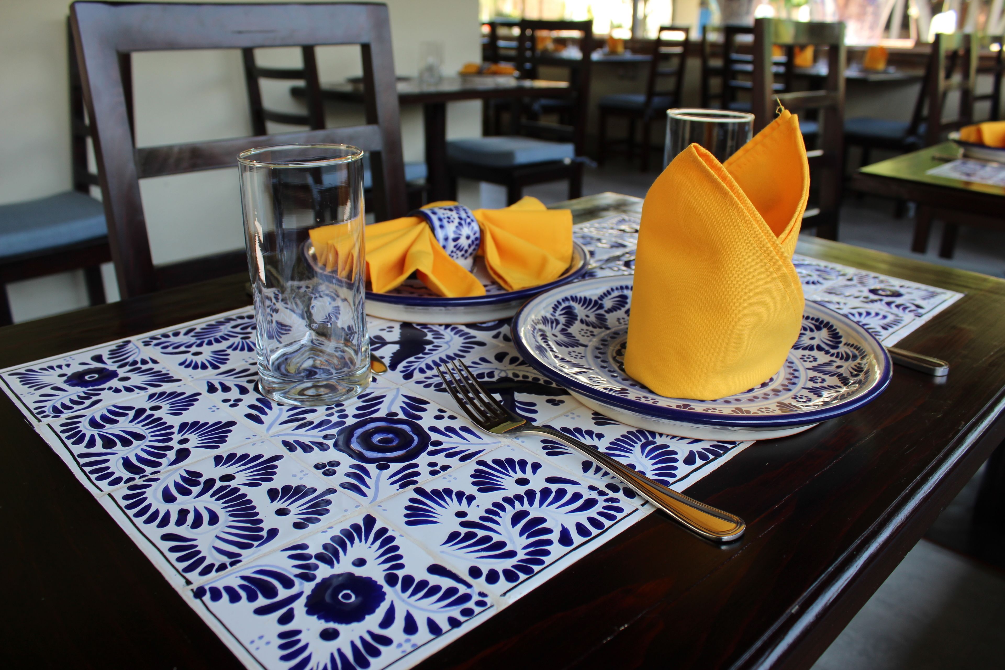 A dining table set with a blue and white patterned tablecloth, a glass of water, a folded yellow napkin, a fork, and a spoon.