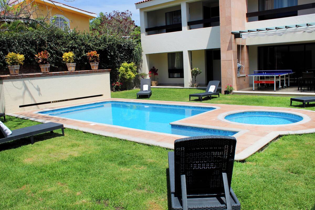 A swimming pool with two hot tubs, surrounded by grass and potted plants.