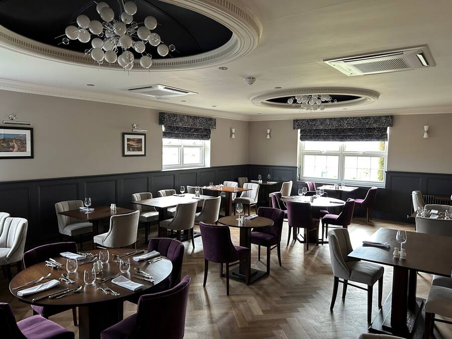 A restaurant with multiple dining tables and chairs, large windows with dark valances, and a chandelier.