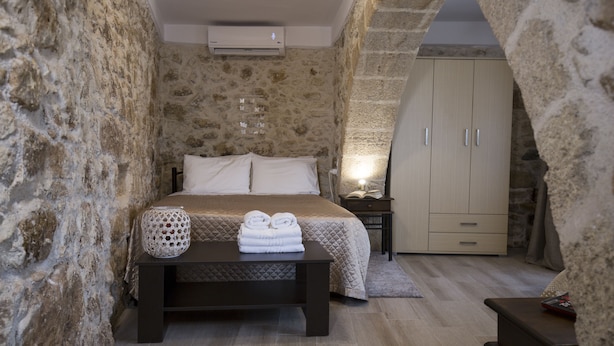 A bedroom with stone walls, a bed with white linens, a bedside table with a lamp, and a wardrobe. - Mylos (Kissamos)