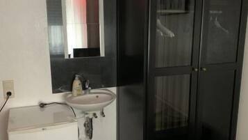Une chambre à coucher de taille modeste, avec une table de chevet blanche, un petit lavabo blanc, une armoire noire et une poubelle.