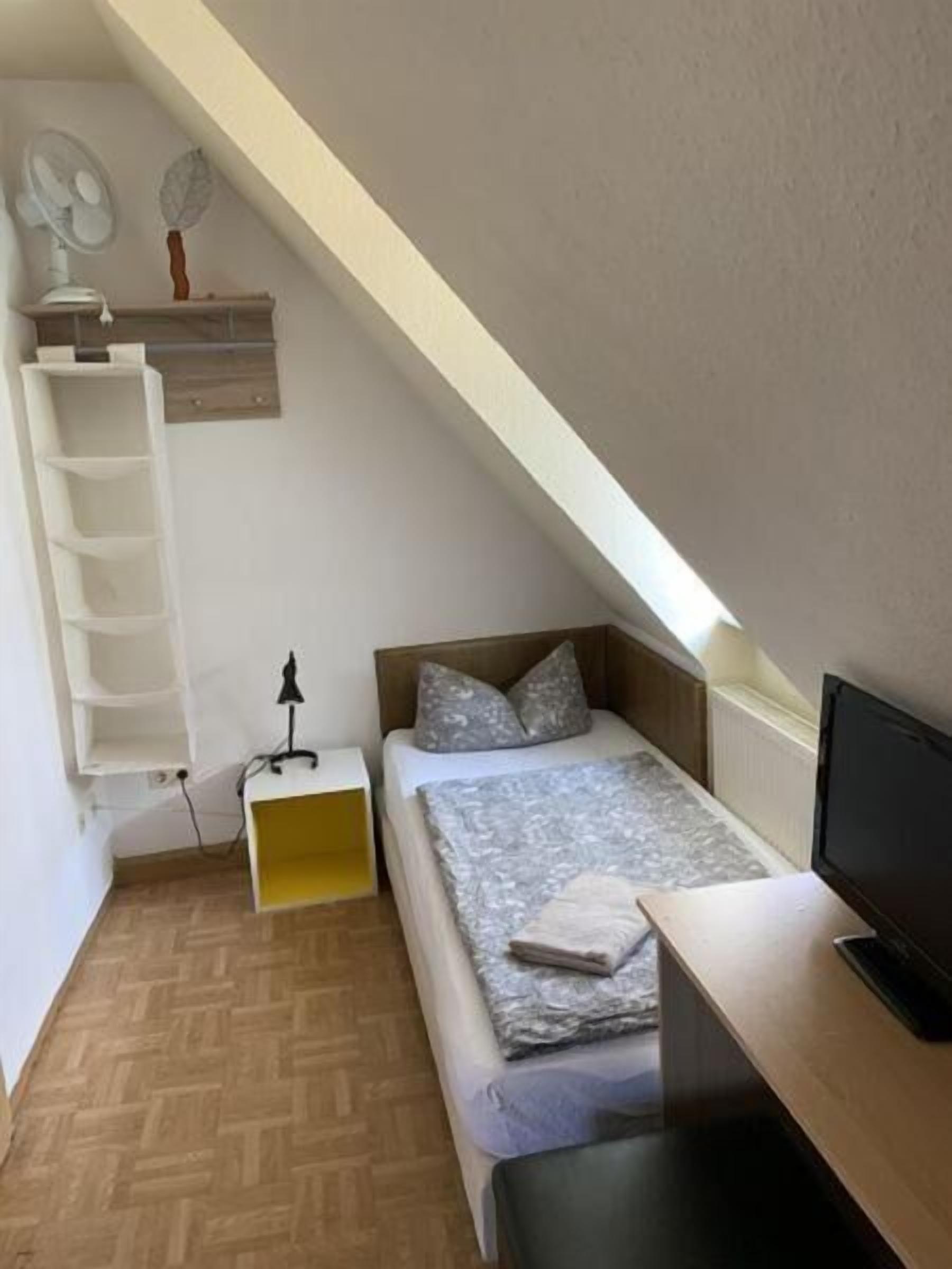 Ein kleines, modernes Schlafzimmer mit schrägem Dach, einem Bett mit grau gemustertem Überwurf, einem Nachttisch mit Lampe und einem Fernseher auf einem Holztisch.