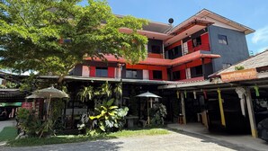 A multi-story hotel with a red and grey facade, a covered parking area, and a variety of plants in the foreground. - Sandee Place Mountain View Chomthong (Chom Thong)