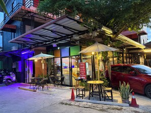 An outdoor cafe with tables and chairs, a red car parked nearby, and a building with a sign in a foreign language. - Sandee Place Mountain View Chomthong (Chom Thong)