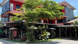 A multi-story building with a red facade, outdoor seating area with umbrellas, and a tree in front. - Sandee Place Mountain View Chomthong (Chom Thong)