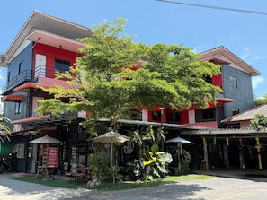 Un edificio de varios pisos con fachada roja, área de estar al aire libre con sombrillas y un árbol en la parte delantera.
