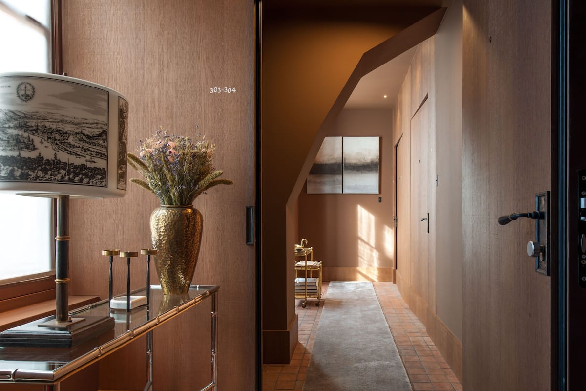 A hallway with a wooden floor, a glass display case with a vase of flowers, and a wall-mounted lamp.