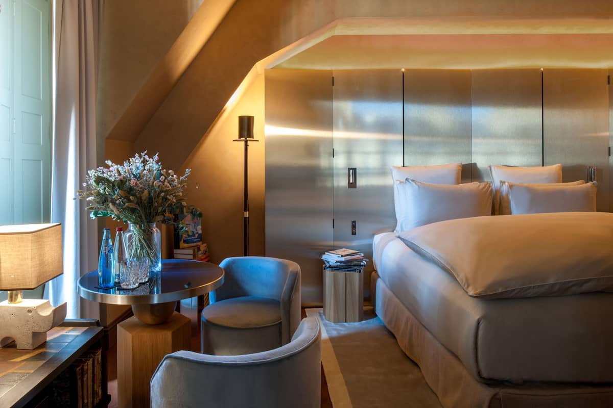 A modern hotel room with a large bed, a sitting area with a table and chairs, a small side table, and a decorative vase with flowers.