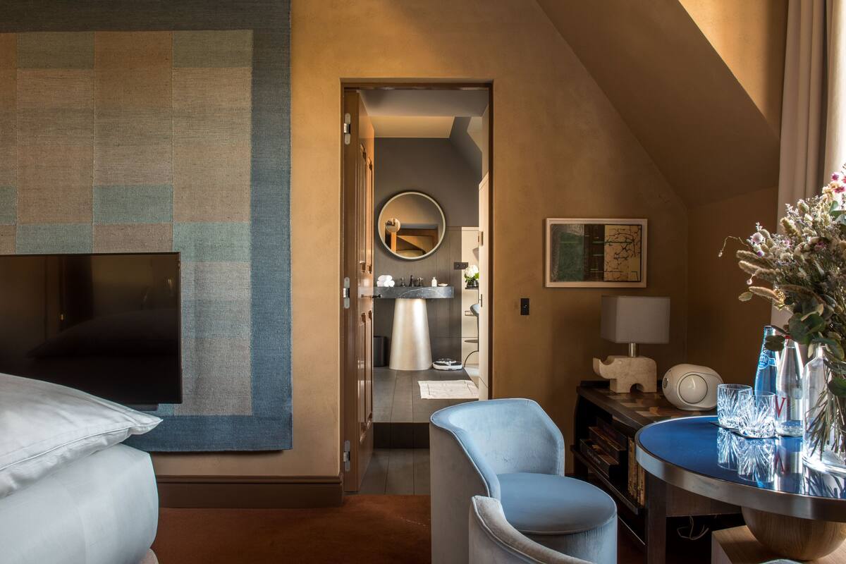 A hotel room with a large bed, a seating area with a chair, a small table with a lamp, a mirror, and a view of the bathroom.