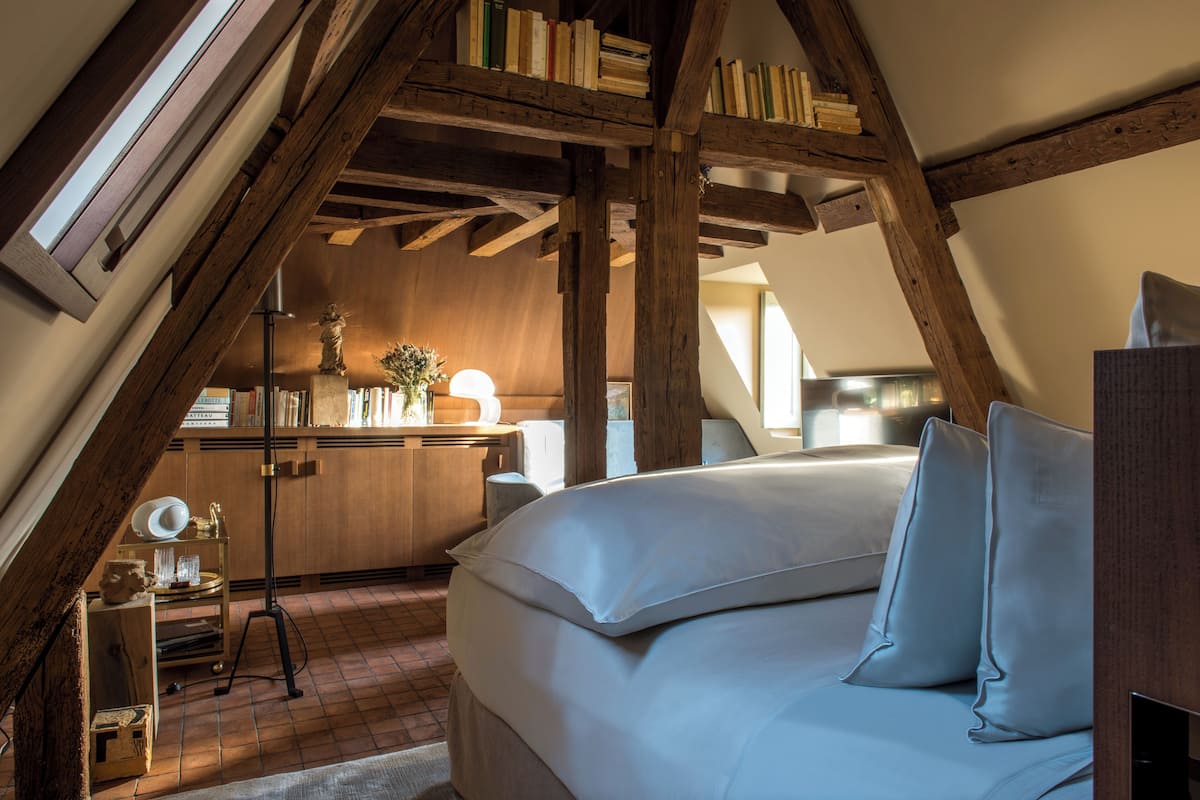 A bedroom with a sloped ceiling, wooden beams, a bed with pillows, a bedside table with a lamp, and a bookshelf filled with books.