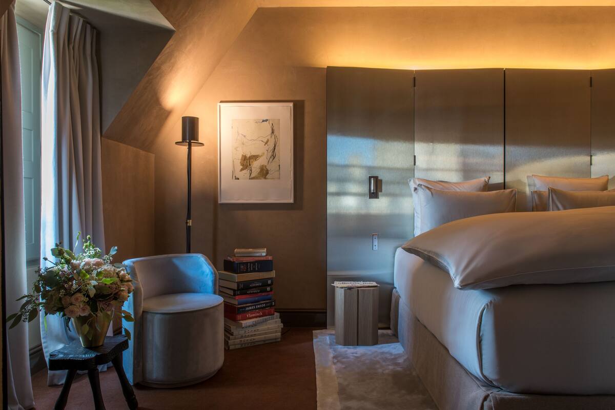 A modern bedroom with a bed, a chair, a small table with books, a vase with flowers, and a framed picture on the wall.