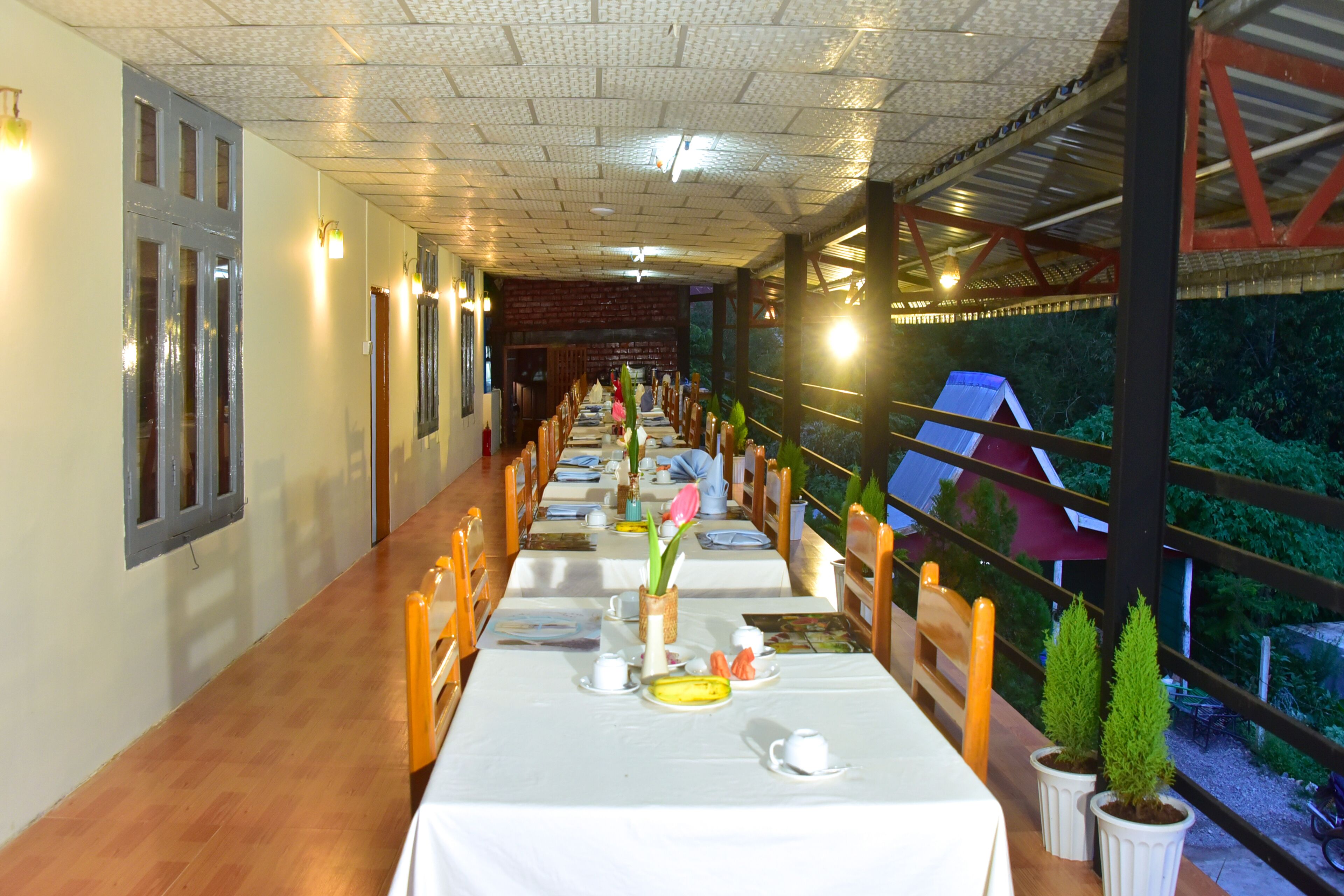 A long dining area with tables set for a meal, wooden chairs, and potted plants.