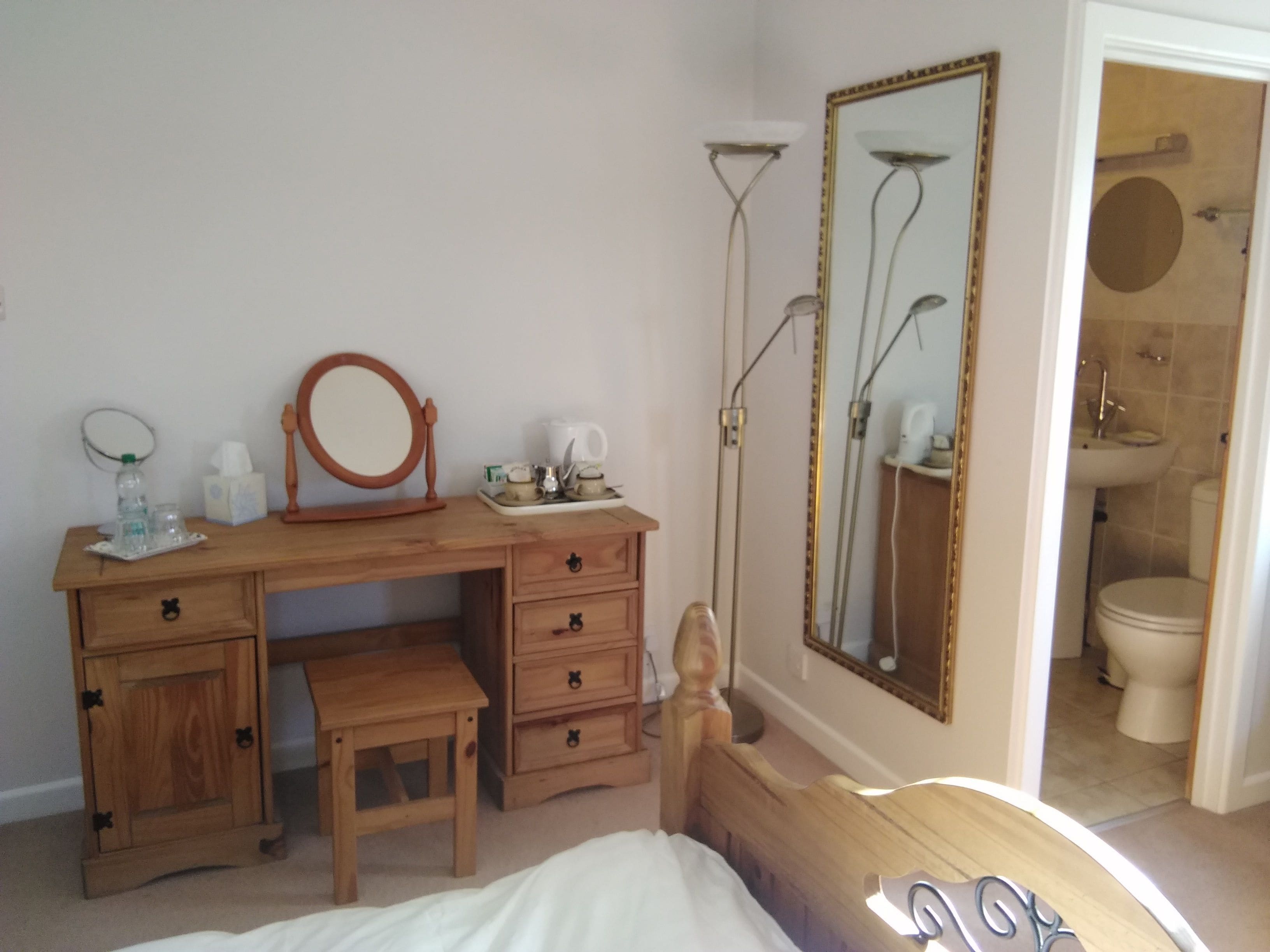 A bedroom with a wooden dresser, a bedside table, a bed, a mirror, and a bathroom visible through an open door.