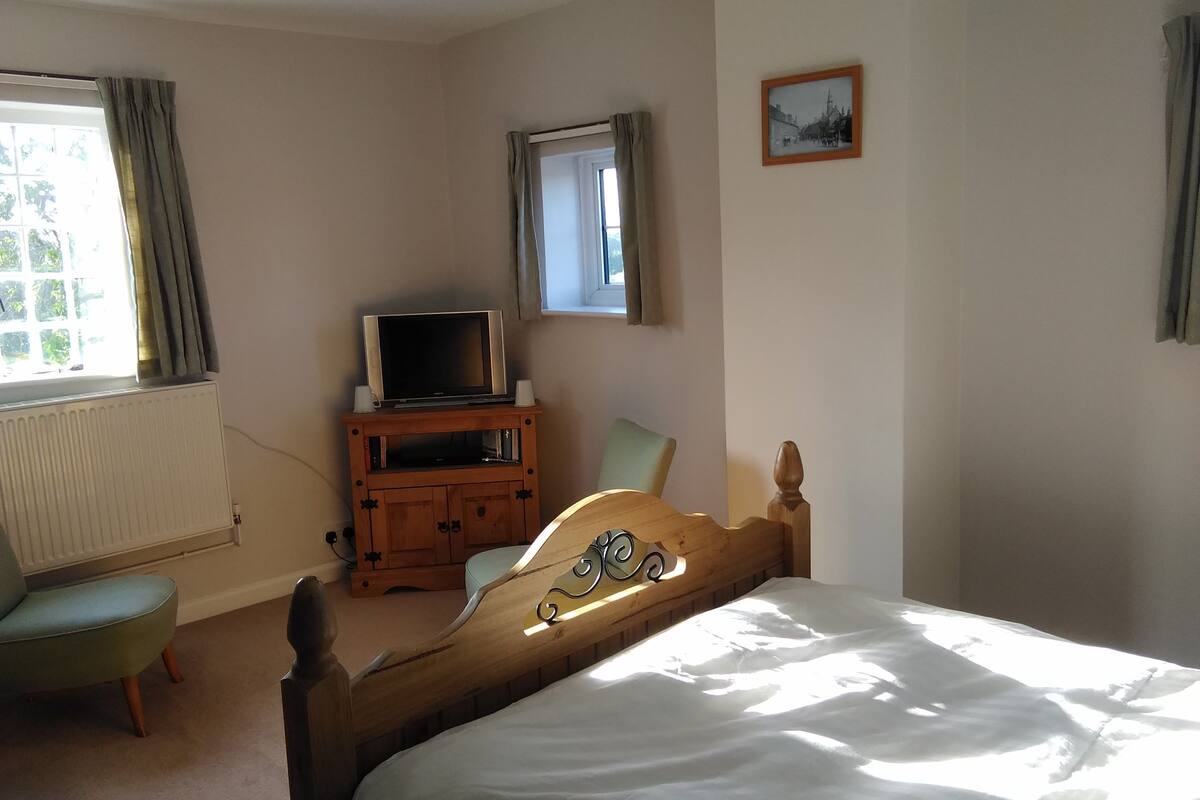 A bedroom with a bed, a wooden dresser, a television on top, a chair, and a window with curtains.