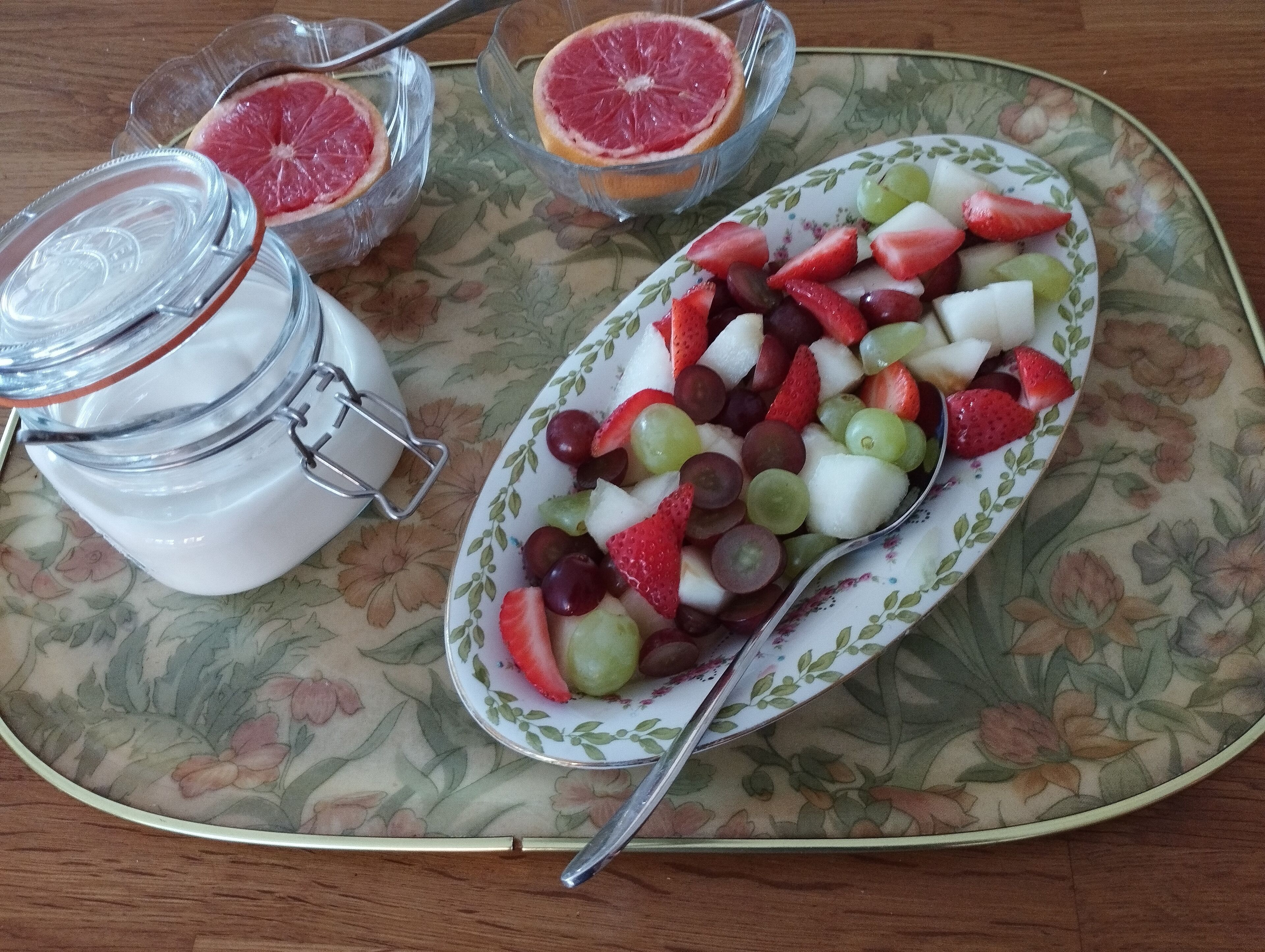 A plate of mixed fruit salad with strawberries, grapes, and melon, accompanied by a glass jar of yogurt and two glasses of grapefruit juice on a floral-patterned tablecloth.