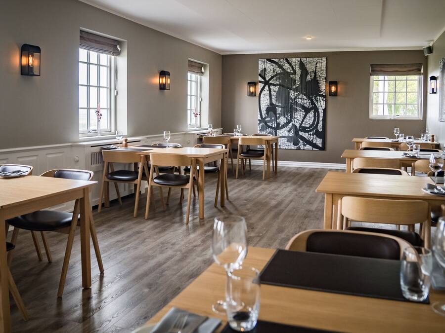 Un restaurant avec des tables et des chaises en bois, une grande peinture abstraite au mur et des fenêtres avec des rideaux.