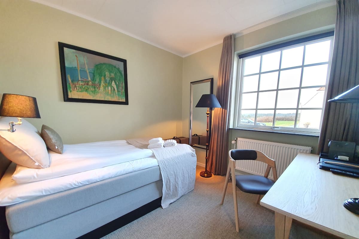 A hotel room with a bed, a desk, a chair, a lamp, a painting on the wall, and a window with a view.