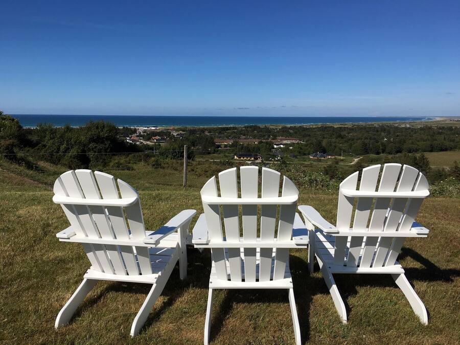 Three white Adirondack chairs facing a scenic view of a coastal town with houses and greenery.