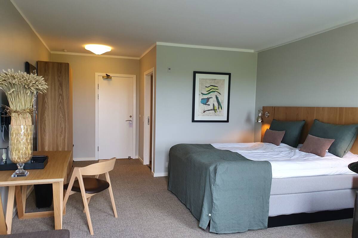 A hotel room with a bed, bedside table, chair, and a desk with a vase of dried flowers.