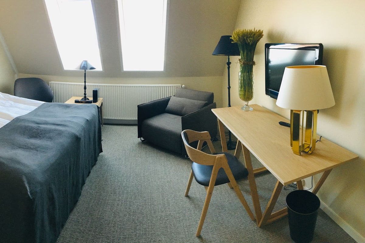 A hotel room with a bed, a desk, a chair, a lamp, and a radiator.