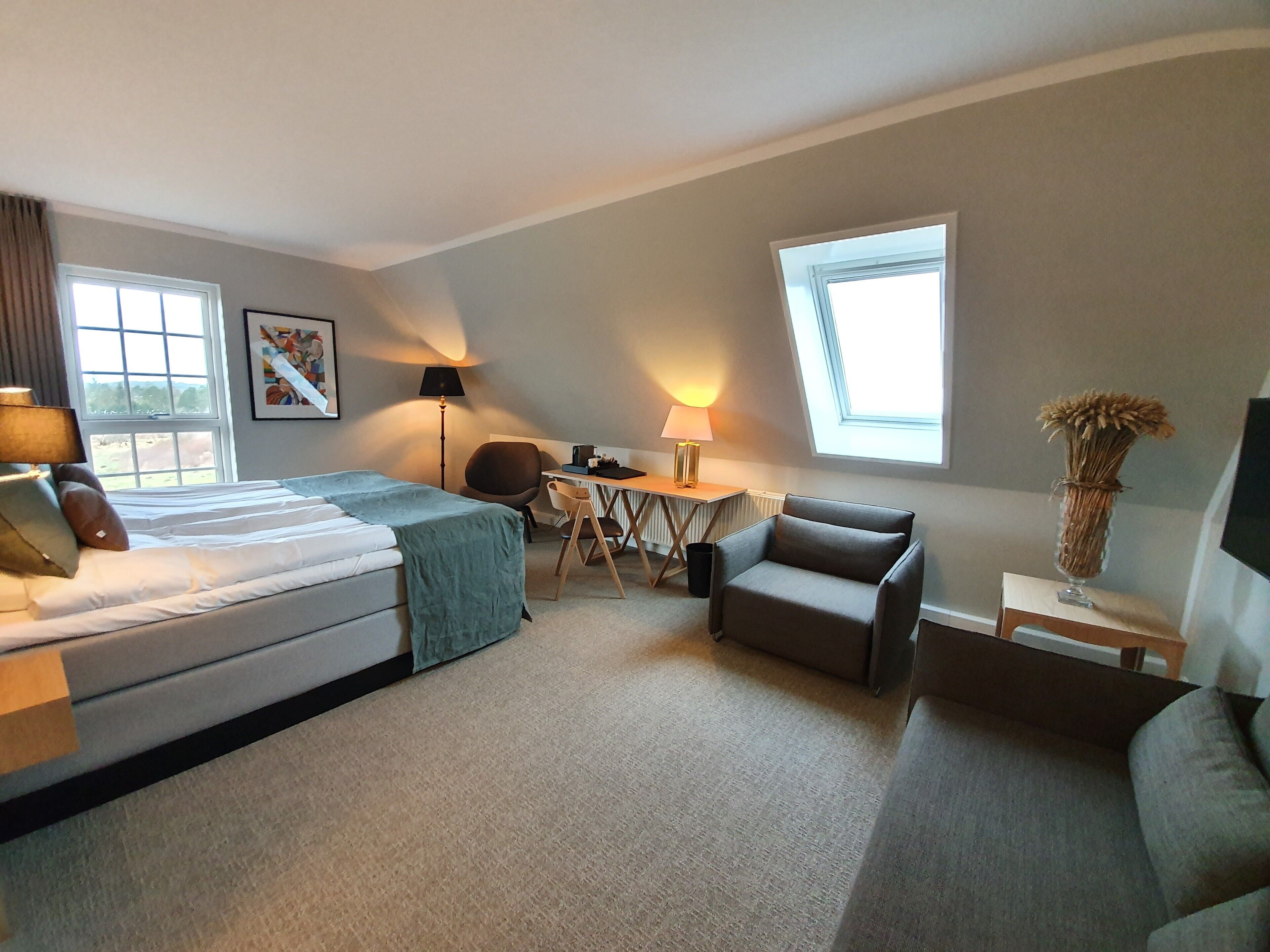 A hotel room with a bed, two chairs, a desk, and a small table. There are windows providing natural light.