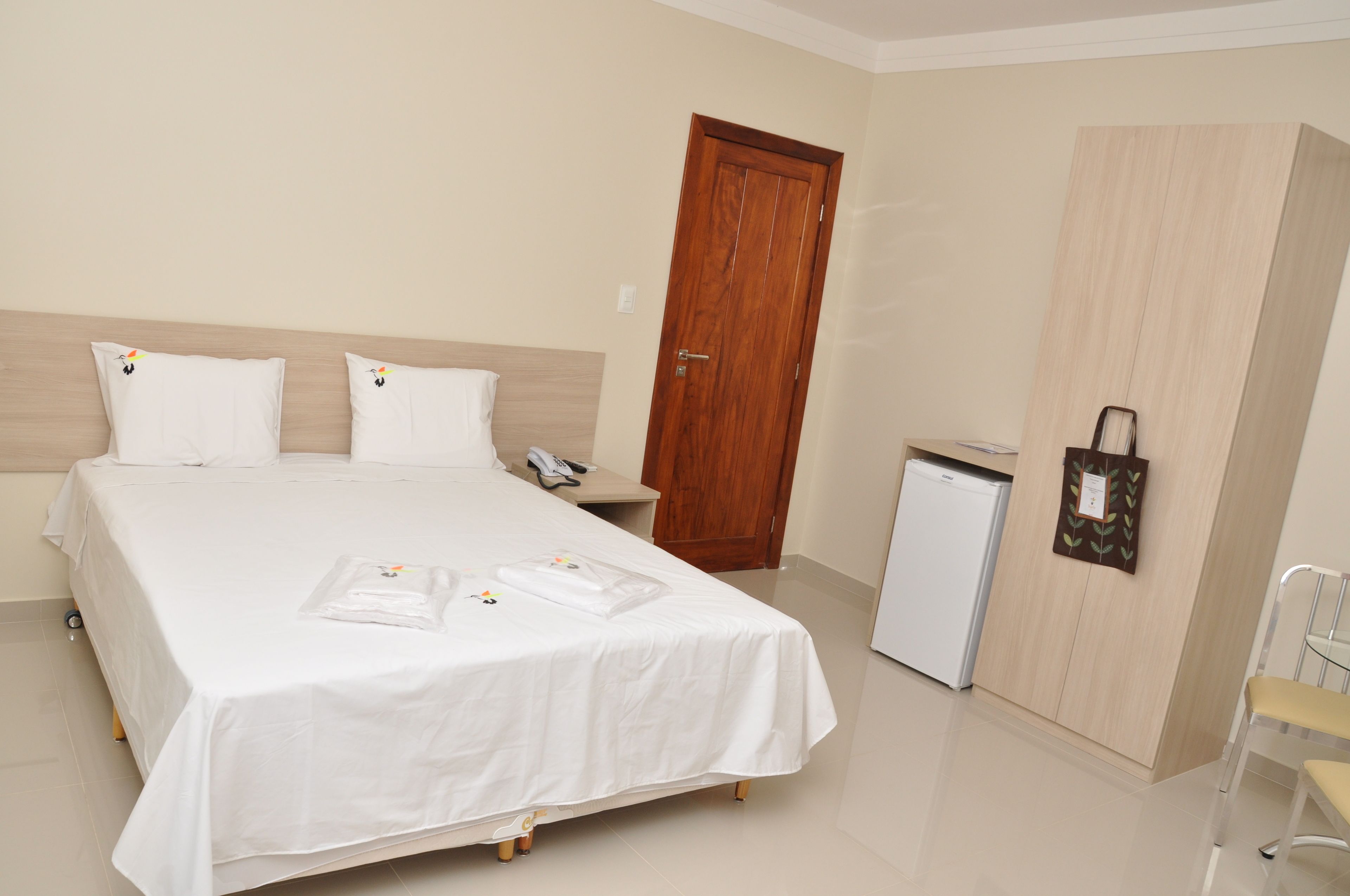 Quarto de hotel com cama, escrivaninha, frigorífico e guarda-roupa de madeira.