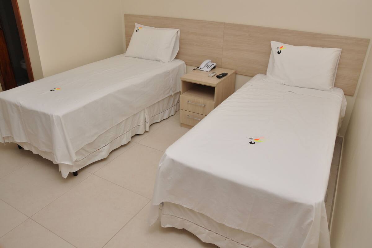Duas camas de solteiro com roupa de cama branca, uma mesa de cabeceira com telefone e uma cômoda pequena.