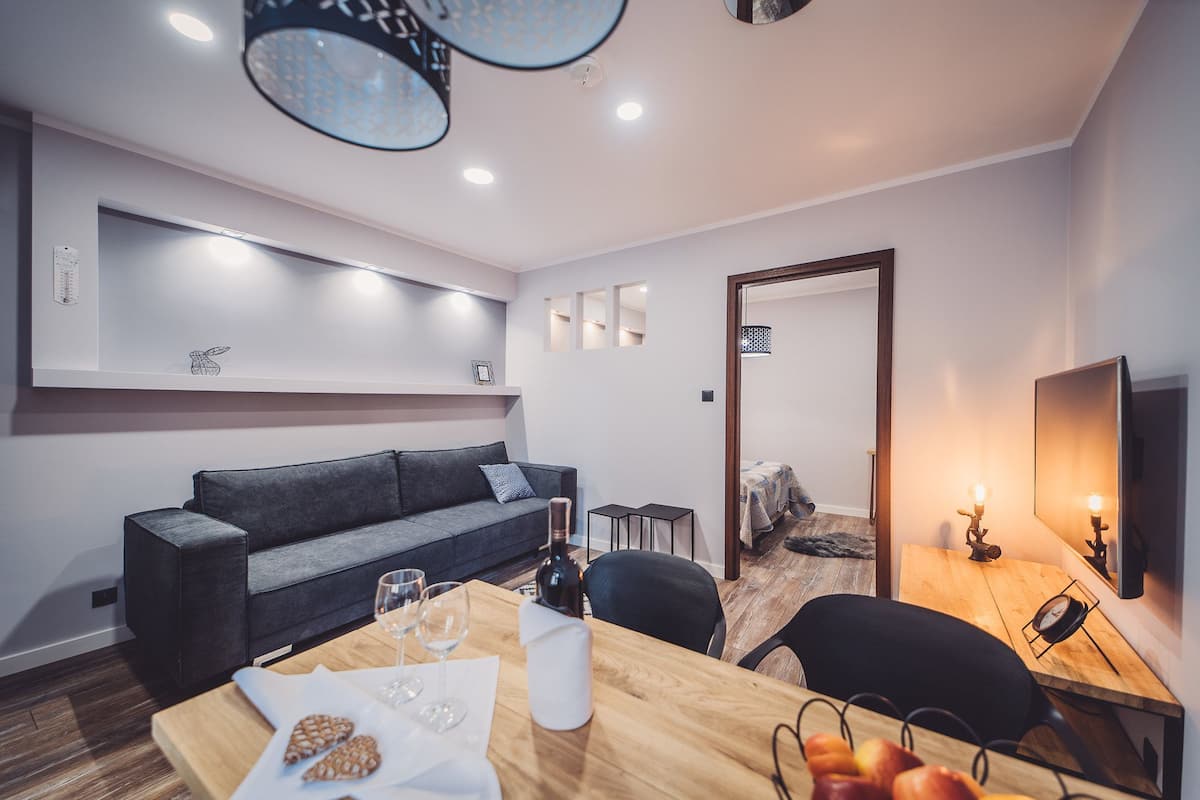 A modern living room with a dark grey sofa, a wooden dining table with wine glasses, and a small kitchen area with a wooden table and chairs.