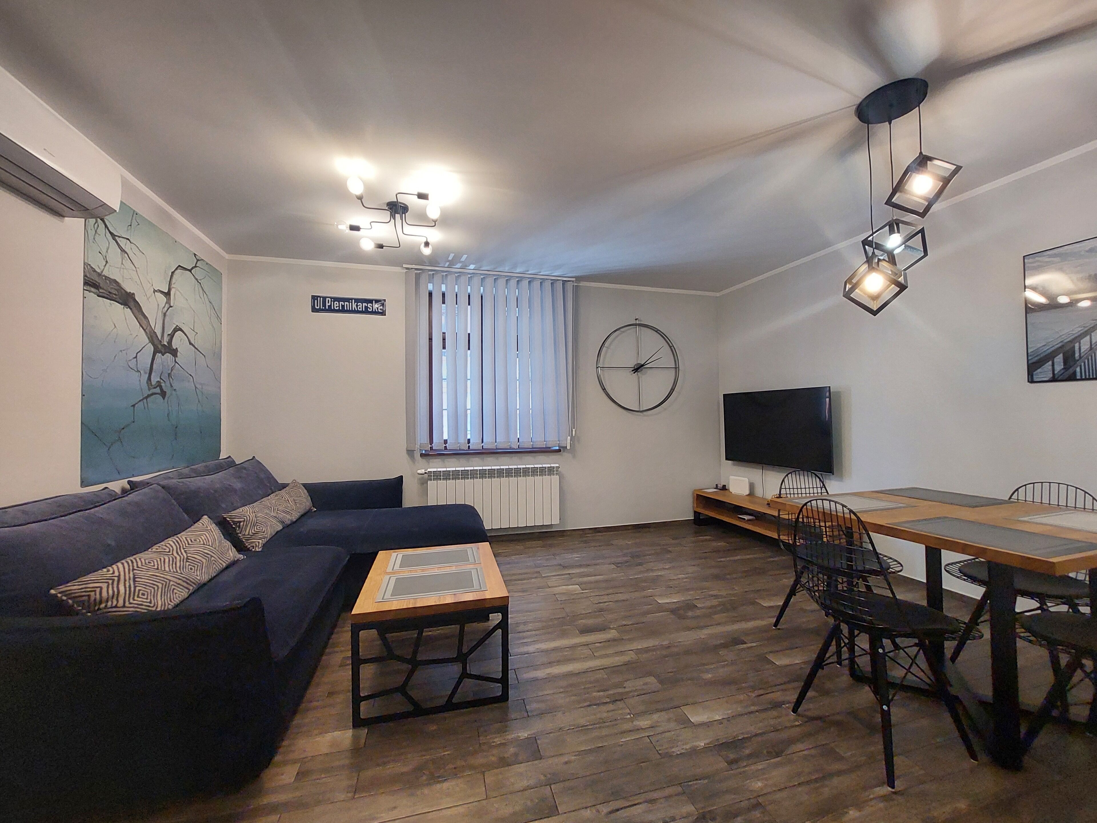 A modern living room with a sofa, a wooden coffee table, a dining table with chairs, a TV, a clock, and a window with blinds.