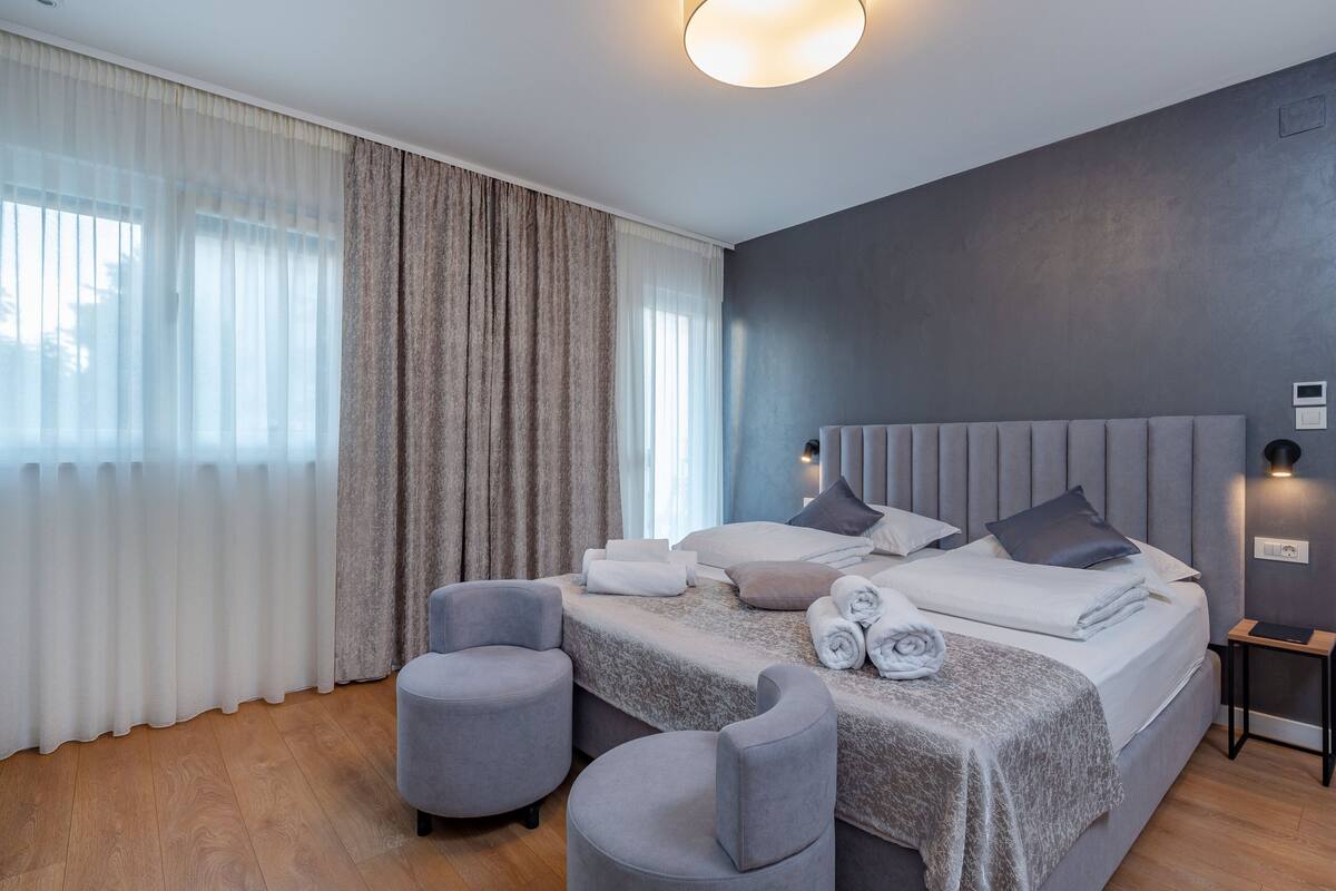 Una habitación de hotel moderna con una cama grande, dos sillones, una mesita de noche y un ventanal con cortinas translúcidas.