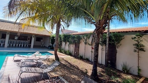 Un área junto a la piscina con sillones de descanso, palmeras y un techo de tejas.
