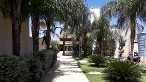 Un camino pavimentado bordeado de palmeras y arbustos, que conduce a un edificio con techo de tejas.