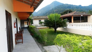 A building with a covered walkway, benches, and a garden with a palm tree. - Pousada Atlantica Recanto do Mar (Caraguatatuba)