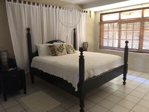 A neatly made bed with a white bedspread and decorative pillows, a wooden headboard, a bedside table with a lamp, and a window with a wooden blind. - Chateau Devaux Cliff Top Villa (Gros Islet)